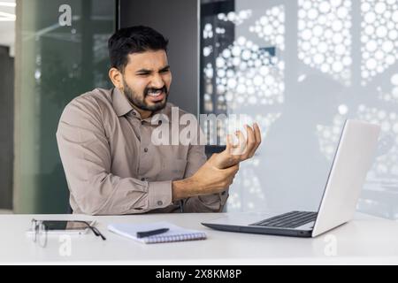 In einer modernen Büroumgebung leidet ein Geschäftsmann bei der Arbeit an einem Laptop unter Schmerzen und Grimmen, wahrscheinlich aufgrund von Belastungen oder Verletzungen durch sich wiederholende Aufgaben, wobei Stress und Unannehmlichkeiten am Arbeitsplatz hervorgehoben werden Stockfoto