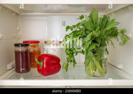 Petersilie, Liebling und Dillblätter in einem Glas mit Wasser, in einem Kühlschrank neben Marmeladengläsern, abstrakt Stockfoto