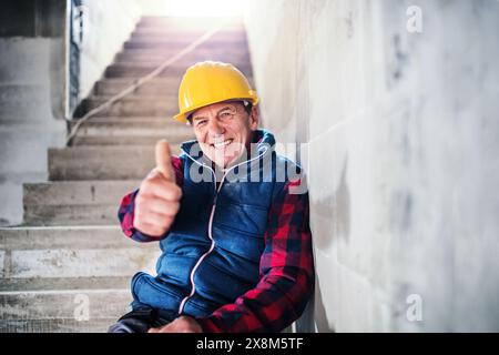 Lächelnder reifer Mann mit Schutzhelm auf der Baustelle, der in die Kamera blickt, auf Betontreppen sitzt. Daumen hoch. Stockfoto