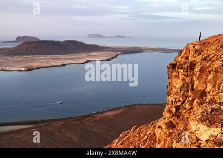 Eine Einzelfigur steht auf einer zerklüfteten Klippe und bestaunt die türkisfarbene Meereslandschaft und die unberührte Wildnis, die sich über die ferne Küste erstreckt. Stockfoto