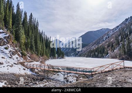 Der malerische Kolsai-See ist eisbedeckt, umgeben von Hügeln mit grünen Fichten und Schnee. Nördlicher Tien Shan, Kolsai-Schlucht, Kasachstan. Frühlingslandschaft mit Stockfoto