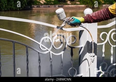 Ein Arbeiter malt einen Metallzaun mit weißer Farbe aus einer Spritzpistole Stockfoto