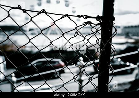 Nahaufnahme eines alten, rostenden Maschendrahtzaugs mit verschwommenen Autos und Gebäuden im Hintergrund an einem bewölkten Tag. Stockfoto