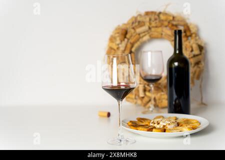 Gläser Rotwein, Weinflasche und gebackener Brie-Käse mit geröstetem Brot auf einem weißen Teller auf einem weißen Tisch, weißem Hintergrund mit einem Kranz aus Weindekor Stockfoto