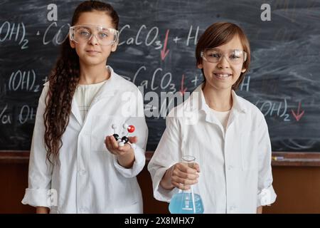 Ethnisch vielfältiges Mädchen und Jungen in Labormänteln und Brillen mit konischer Flasche und chemischem Strukturmodell, das an der Tafel steht Stockfoto