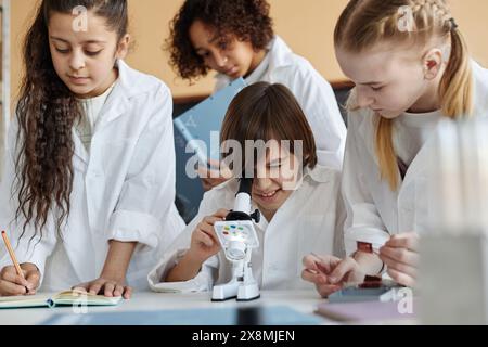 Gruppe ethnisch unterschiedlicher Mittelschüler, die Labormäntel tragen, experimentieren im Chemiekurs mit Mikroskop Stockfoto