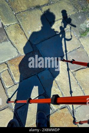 Außenschatten des Ganzkörperfotografen, der Kamera und des orangen Reisestativs stehen auf gelben Betonsteinpflastern. Die Zeit für Fotoaufnahmen ist nahe. Stockfoto