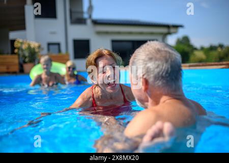 Ein paar fröhliche Senioren rasieren Spaß im Pool mit Freunden, die springen, schwimmen und auf Wagen liegen. Ältere Freunde verbrachten einen heißen Tag am Swimmingpool Stockfoto