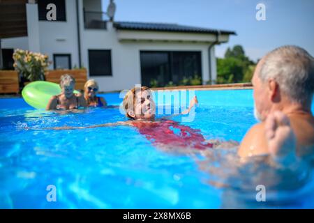 Ein paar fröhliche Senioren rasieren Spaß im Pool mit Freunden, die springen, schwimmen und auf Wagen liegen. Ältere Freunde verbrachten einen heißen Tag am Swimmingpool Stockfoto