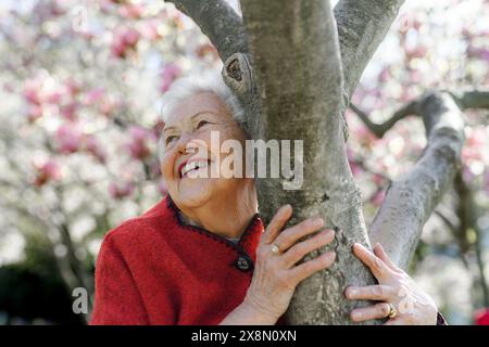 Porträt einer älteren Frau, die am Magnolienbaum steht, im Park, einen entspannenden Moment hat. Großmutter genießt warmes Frühlingswetter. Stockfoto