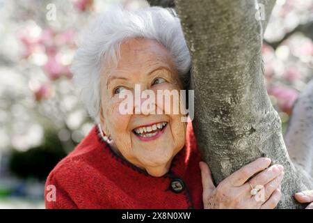 Porträt einer älteren Frau, die am Magnolienbaum steht, im Park, einen entspannenden Moment hat. Großmutter genießt warmes Frühlingswetter. Stockfoto