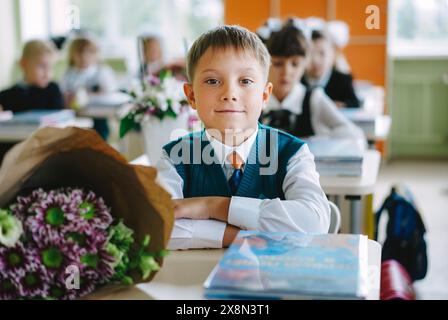 Niedlicher russischer Junge, der am 1. september in einem Klassenzimmer mit neuem, modernem Roller am Schreibtisch sitzt Stockfoto