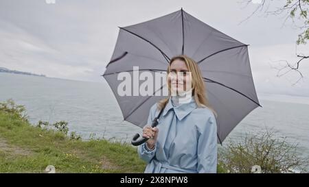 Junge lächelnde Frau mit Augen voller Liebe, die einen grauen Regenschirm spinnt. Stock Clip. Mädchen im Mantel an der Meeresküste im Frühling, das mit jemandem spricht. Stockfoto