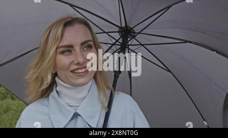 Junge blonde Frau mit grauem Schirm draußen auf grüner Wiese. Stock Clip. Regenwetter mit Regenschirm. Stockfoto