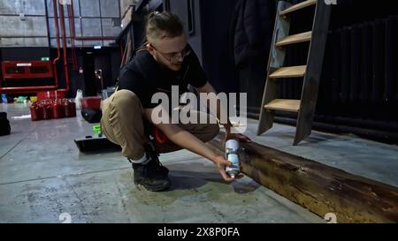 Spritzlackierung von Holzbalken in einem Konzertsaal. Medien. Junger Mann, der Holzbalken mit schwarzer Farbe dekoriert. Stockfoto