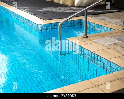 Haltegriff aus Chrom-Metall, klare Wasseroberfläche am Pool mit Treppenbereich. Abstrakte Poolstruktur, blaue Gitterfliesen mit Unterwassermuster, n Stockfoto