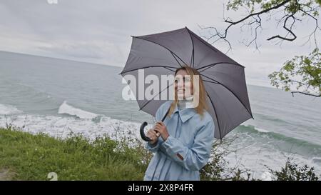 Junge lächelnde Frau mit Augen voller Liebe, die einen grauen Regenschirm spinnt. Stock Clip. Mädchen im Mantel an der Meeresküste im Frühling, das mit jemandem spricht. Stockfoto