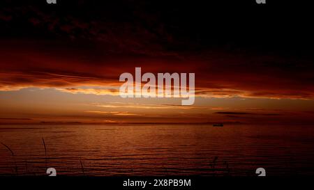 Märchenhafter atemberaubender Sonnenuntergang über dem plätschernden Fluss mit einem schwimmenden Boot. Clip. Dunkelroter und oranger Himmel, schwere Wolken über Wasser. Stockfoto