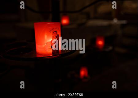 In einer schwach beleuchteten Kirche in Carcassonne flackert eine rote Kerze mit einem heiligen, während andere sanft leuchtende Kerzen im Hintergrund stehen. Stockfoto