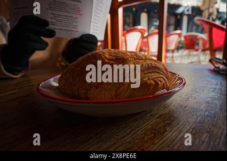 Goldenes Croissant sitzt auf einem weißen Teller mit rotem Rand; schwarze Hände schmiegen ein Menü im Hintergrund, alles auf einem Holztisch. Stockfoto