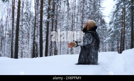 Glücklicher junger Mann im märchenhaften Winterwald betet zu Gott, der auf den Knien steht. Medien. Religionsbegriff und das Gefühl der Einheit mit der Natur. Stockfoto