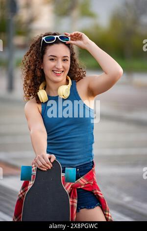 Eine fröhliche junge Frau mit lockigem Haar, Sonnenbrille und Kopfhörer, hält ein Skateboard in einem urbanen Park. Stockfoto