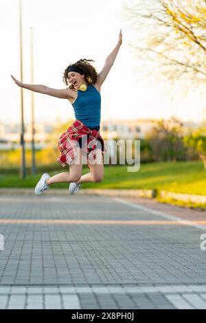 Glückliche junge Frau in blauem Tanktop und rotem kariertem Hemd, fröhlich mit Kopfhörern hüpft, Arme weit ausgebreitet, in einem sonnigen Stadtpark bei Sonnenuntergang. Stockfoto