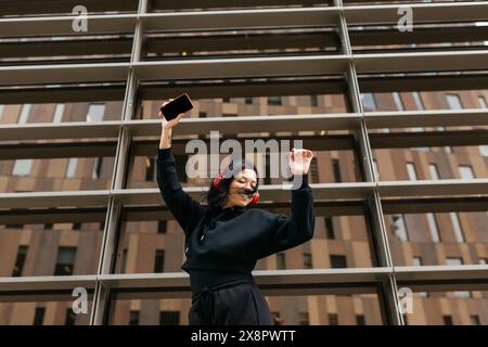 Eine junge Frau in einem schwarzen Hoodie, die mit Kopfhörern und einem Telefon in der Hand vor dem Hintergrund eines modernen Gebäudes tanzt. Stockfoto