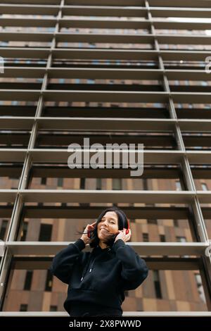 Eine junge Frau in einem schwarzen Hoodie, die mit Kopfhörern und einem Telefon in der Hand vor dem Hintergrund eines modernen Gebäudes tanzt. Stockfoto