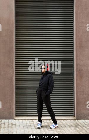 Porträt einer jungen Frau in schwarzem Hoodie, roten Kopfhörern und Sonnenbrille, die Musik in coolem, urbanem Street-Style hört. Stockfoto