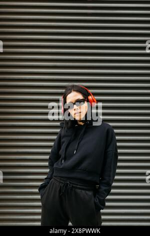 Porträt einer jungen Frau in schwarzem Hoodie, roten Kopfhörern und Sonnenbrille, die Musik in coolem, urbanem Street-Style hört. Stockfoto