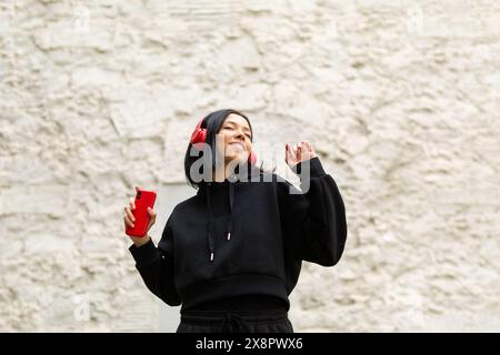 Das Porträt einer jungen Frau in schwarzer Kleidung und Kopfhörern singt fröhlich zur Musik mit, mit einem hellen und fröhlichen Ausdruck auf ihrem Gesicht. Stockfoto