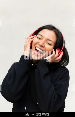 Das Porträt einer jungen Frau in schwarzer Kleidung und Kopfhörern singt fröhlich zur Musik mit, mit einem hellen und fröhlichen Ausdruck auf ihrem Gesicht. Stockfoto