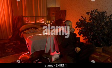Junger bärtiger blonder Mann, der auf dem Boden neben seinem Bett sitzt. Medien. Mann schüttelt den Kopf, während er Musik im Schlafzimmer mit Neonlampen hört. Stockfoto