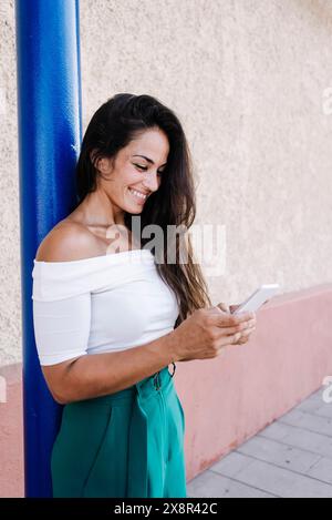 Eine junge Frau mit langen Haaren, die sich an die Wand lehnt und auf das Handy blickt Stockfoto