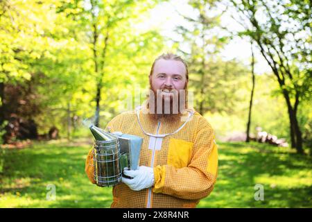Lächelnder Mann mit rotem Bart im Imkeranzug, der einen Raucher hält. Stockfoto