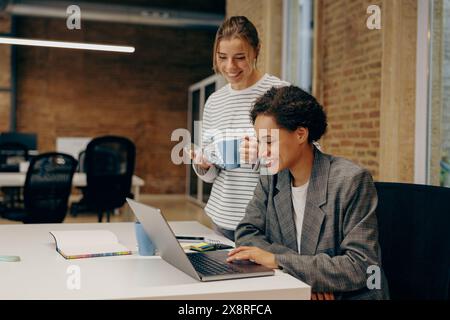 Zwei lächelnde weibliche Kollegen arbeiten gemeinsam an einem Projekt und verwenden im Büro einen Laptop Stockfoto