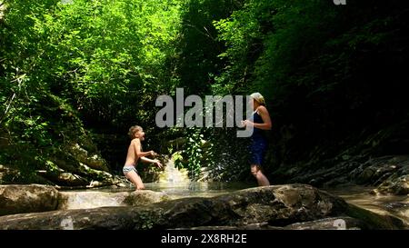 Mutter mit Jungen, die Wasserspritzer im Fluss und im Dschungel im Hintergrund machen. Kreativ. Erkunden Sie wunderschöne Orte in der Natur. Stockfoto