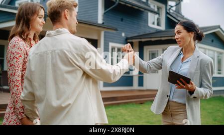 Glücklicher Immobilienmakler gratuliert jungen Hauseigentümern zum Erwerb neuer Häuser. Fröhliches Paar, das der Geschäftsfrau die Hand schüttelt und Schlüssel von ihrem Eigentum erhält. Stockfoto