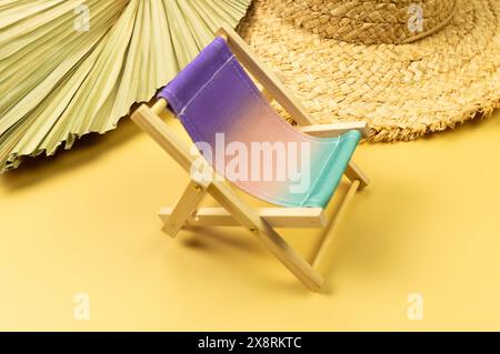 Farbenfroher Strandstuhl, Strohhut, getrocknetes Palmblatt auf gelbem Hintergrund. Sommer-, Urlaubs- und Strandkonzept. Kreative Komposition, Kopierbereich. Stockfoto