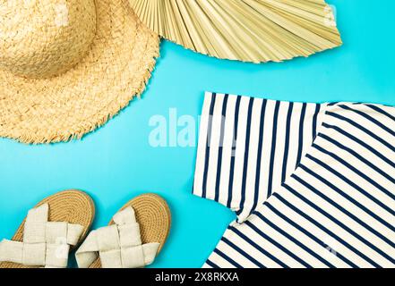 Blick von oben auf Strohhut, Hausschuhe, getrocknete Palmblätter und gestreiftes T-Shirt auf blauem Hintergrund. Sommermode, Urlaubs- und Strandtapete. Kopierbereich, fla Stockfoto