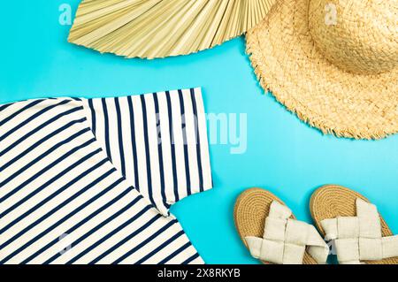 Blick von oben auf Strohhut, Hausschuhe, getrocknete Palmblätter und gestreiftes T-Shirt auf blauem Hintergrund. Sommermode, Urlaubs- und Strandtapete. Kopierbereich, fla Stockfoto