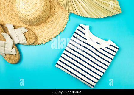 Blick von oben auf Strohhut, Hausschuhe, getrocknete Palmblätter und gestreiftes T-Shirt auf blauem Hintergrund. Sommermode, Urlaubs- und Strandtapete. Kopierbereich, fla Stockfoto