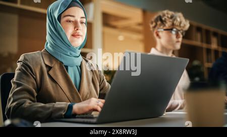 Gruppe verschiedener junger kreativer Kollegen, die nebeneinander sitzen und in Modern Office an Laptops arbeiten. Muslimische Frau lächelt, nachdem sie eine Aufgabe erfolgreich beendet hat. Office-Team-Arbeit Stockfoto