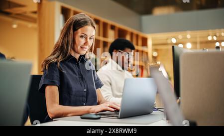 Multiethnische Gruppe kreativer Kollegen, die tagsüber in einem modernen hellen Büro an Laptops arbeiten. Eine weiße Frau lächelt glücklich, nachdem sie ein Arbeitsprojekt erfolgreich beendet hat. Stockfoto