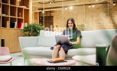 Porträt einer kaukasischen kreativen jungen Frau, die in einem hellen, ungezwungenen Büro an einem Laptop arbeitet. Weibliche Kundenkoordinatorin Lächelt, Wenn Sie Positives Feedback Von Kunden Erhält. Mitarbeiter gehen im Hintergrund Stockfoto