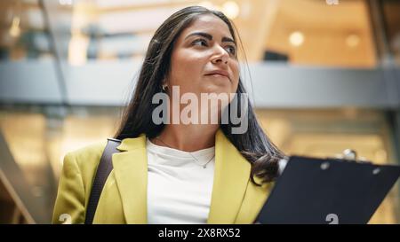 Porträt einer selbstbewussten hispanischen Frau, die ein Firmenbüro überquert, lächelnd und selbstbewusst ist. Eine erfolgreiche Führungskraft geht morgens zu ihrem Schreibtisch und überprüft ihre Aufgaben. Stockfoto