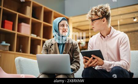 Zwei junge kreative Kollegen diskutieren das Arbeitsprojekt und zeigen auf den Laptop-Bildschirm im Büro. Kaukasischer männlicher Supervisor verwendet ein Tablet und konsultiert die muslimische weibliche Teamleiterin. Stockfoto