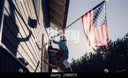 Stolzer Patriotischer Vater hält seine junge Tochter auf den Schultern und hilft ihr, die Flagge der Vereinigten Staaten von Amerika auf die Mauer ihres Hauses zu setzen, um einen Nationalfeiertag zu feiern. Sonniger Tag in den USA. Stockfoto
