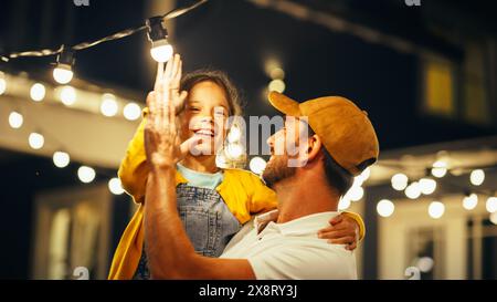 Stolzer, gutaussehender Vater hilft seiner kleinen, schönen Tochter, eine Glühbirne in der Hinterhof-Installation von Fairy Lights zu Hause zu wechseln. Vater und Tochter High Five und feiern erfolgreiche Fix. Stockfoto